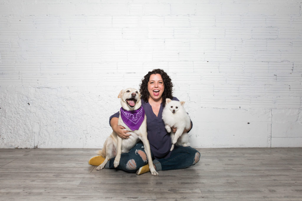 Founder of Pet Boss Nation sitting on wooden floor holding two white dogs, one with a purple bandana