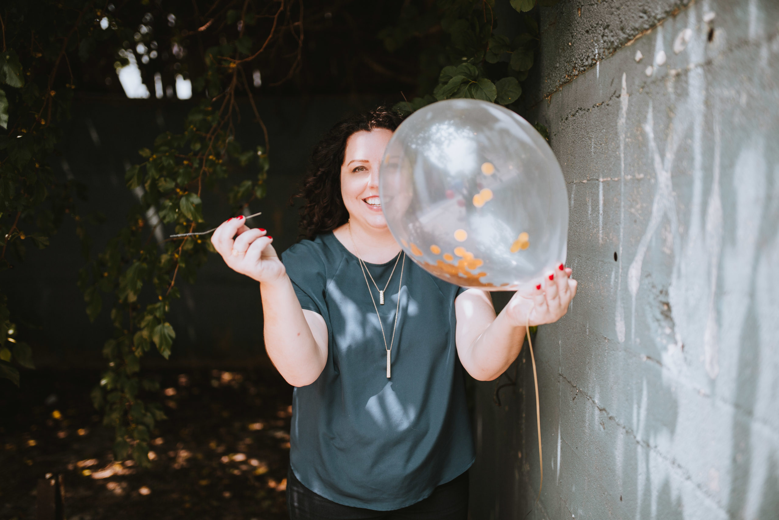 Candace D'Agnolo standing next to a brick wall in an overgrown area holding a balloon in one hand and a needle in the other