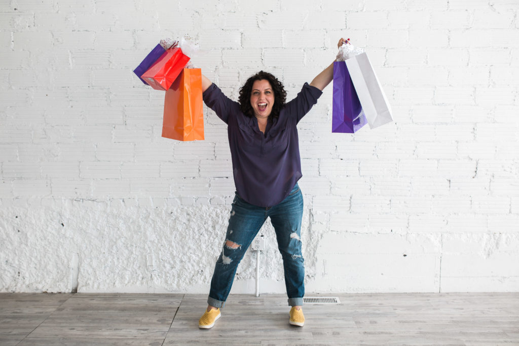 Candace D'Agnolo standing in front of a white brick wall holding up red, orange, purple, and white shopping bags